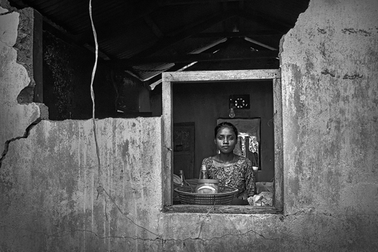 A tea garden labourer from Hantupara, Assam, stands in the window of her broken house, which was damaged by an elephant, as she awaits acknowledgement of the conflict from the tea garden authority.