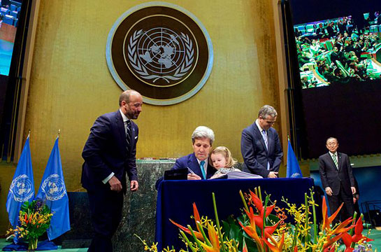 U.S. Secretary of State John Kerry, with his two-year-old granddaughter Isabelle Dobbs-Higginson on his lap and United Nations Secretary-General Ban ki-Moon looking on, signs the COP21 Climate Change Agreement on behalf of the United States during a ceremony on Earth Day, April 22, 2016, at the U.N. General Assembly Hall in New York, N.Y. [State Department photo/ Public Domain]